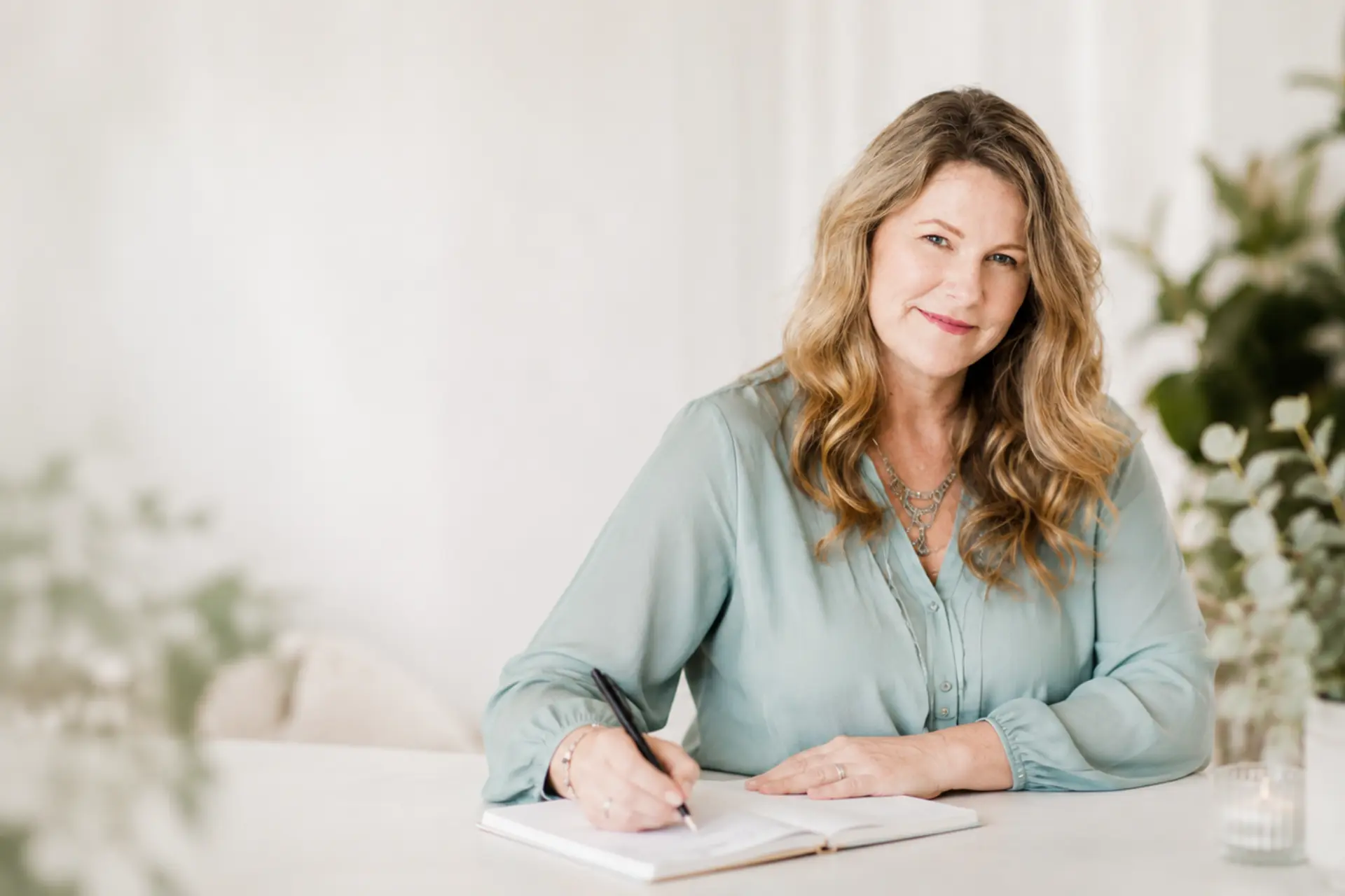 Lori Satterthwaite of Lola Media Design writing in a notebook in her warm, plant-filled Matakana studio